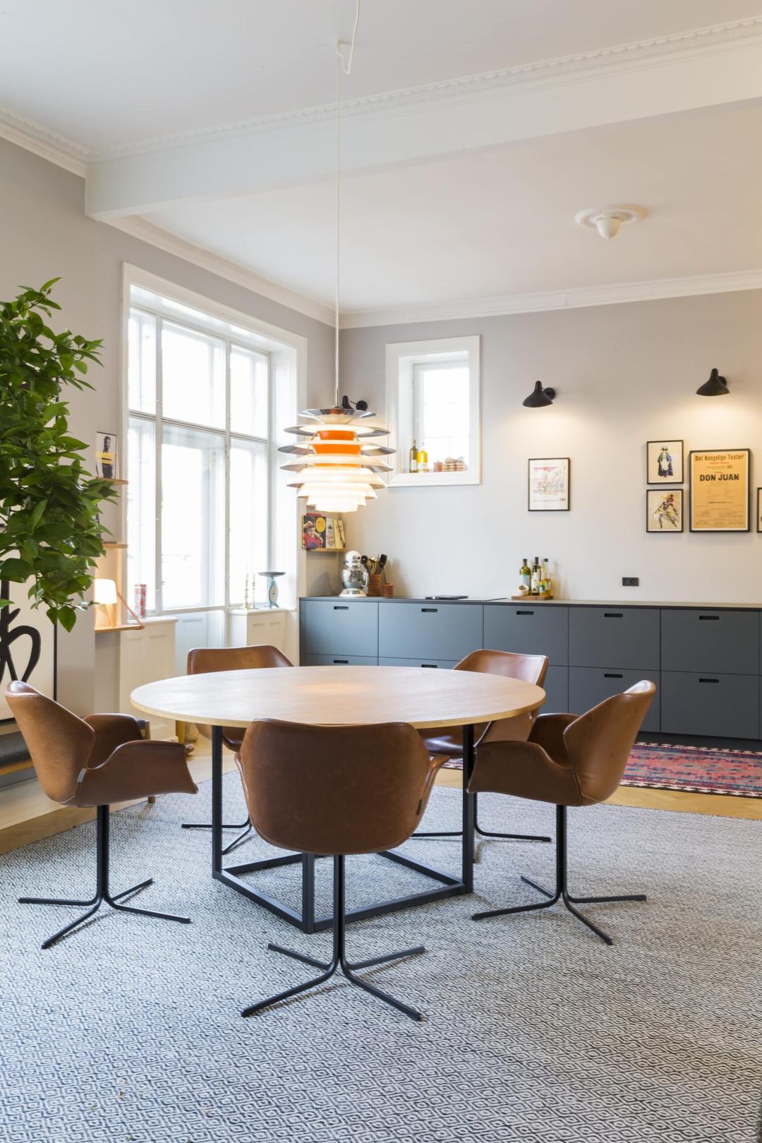 Lys spisestue med rundt træbord, brune læderstole, moderne pendel og skænk med kunst på væggen