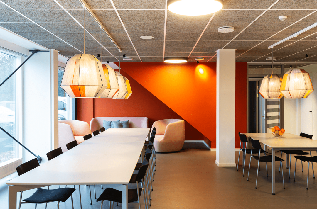 Spiseområde med lange borde, pendellamper og rød væg, der giver moderne og dynamisk indretning.