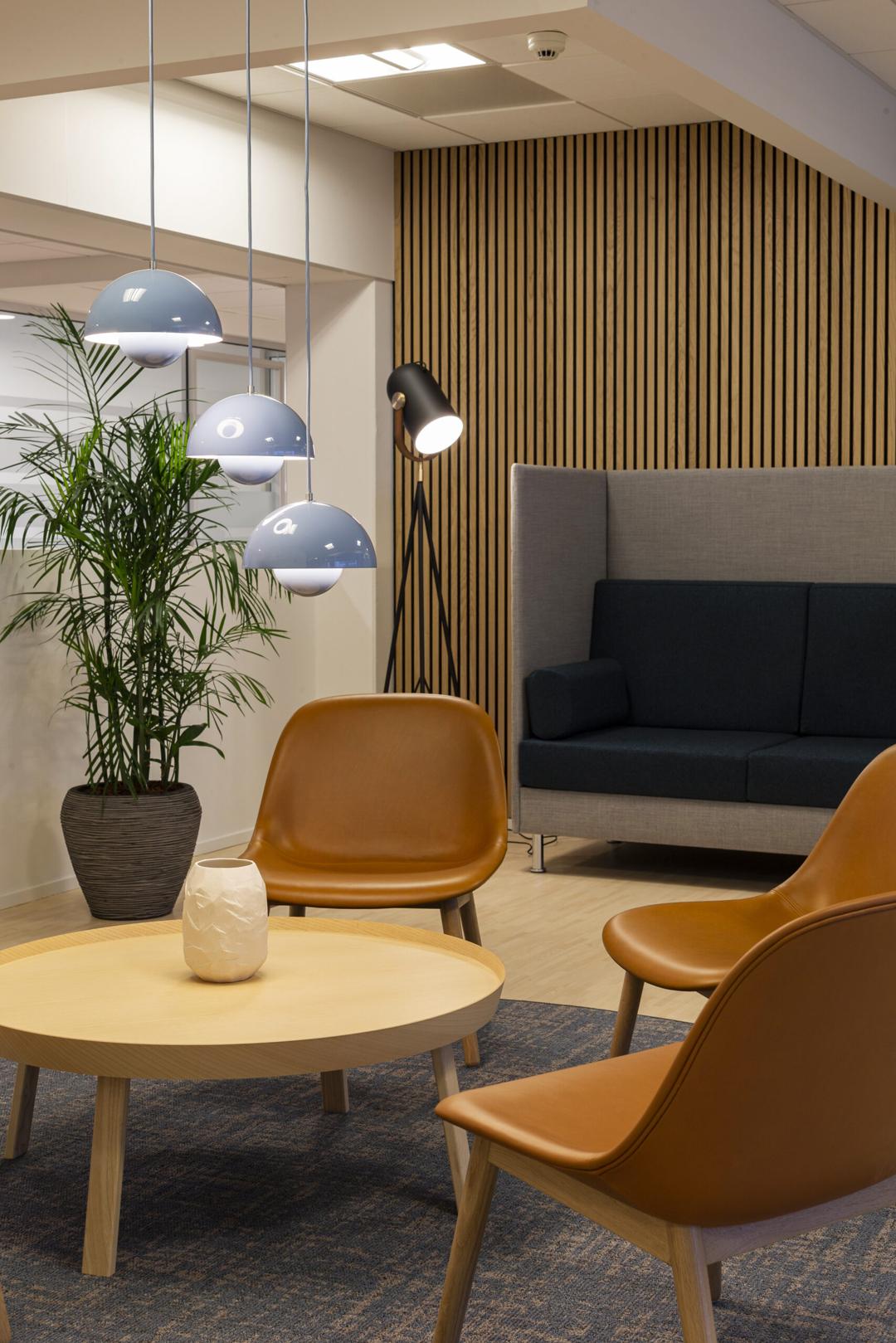 Et rundt bord, orange stole, plads til samtale og afslapning på kontoret, elegant indretning, grå hængelamper og en høj gulvlampe i hjørnet.