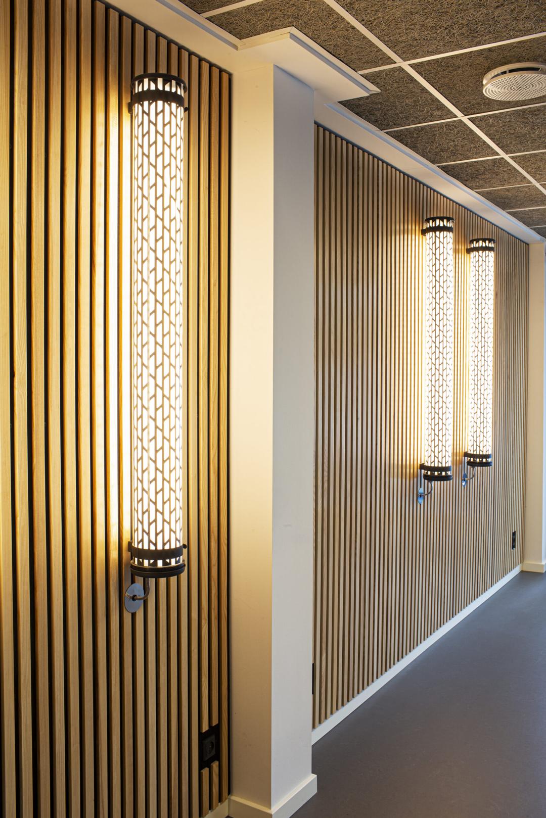Gangareal med moderne vægbelysning monteret på en væg med lodrette trælameller, der giver et varmt og stilrent udtryk.