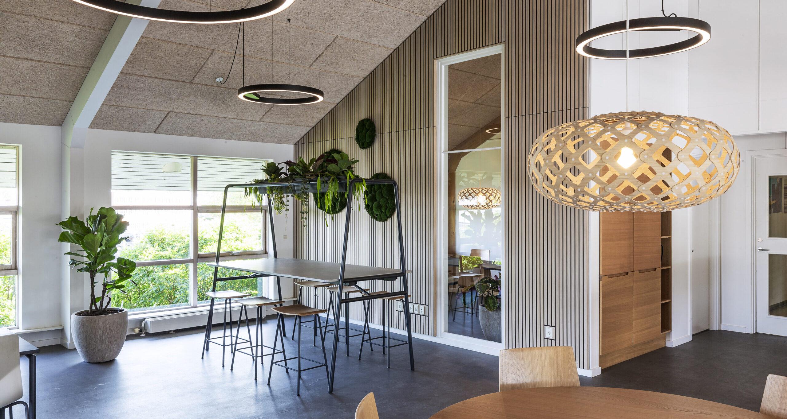 Moderne kontorindretning med højbord, akustikloft, trælameller, grønne planter og designerlamper i lyst skandinavisk kontormiljø.