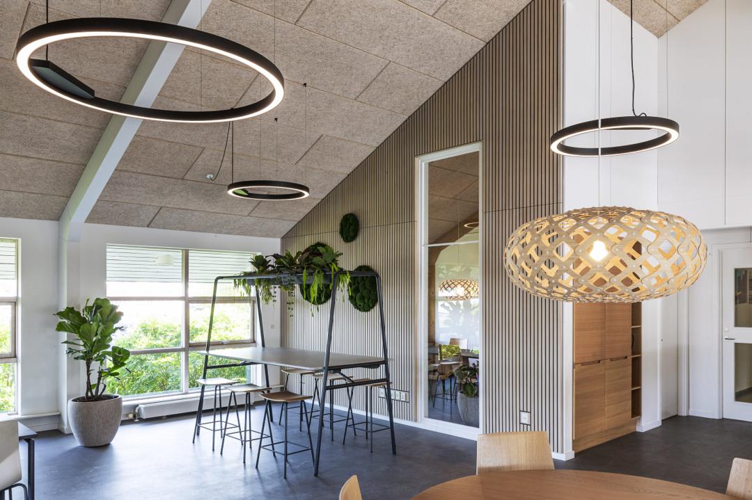 Moderne kontorindretning med højbord, akustikloft, trælameller, grønne planter og designerlamper i lyst skandinavisk kontormiljø.