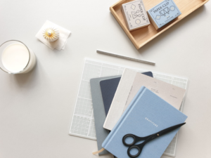 Dekorative kontorartikler og notesbøger i blå nuancer på hvidt bord med duftlys og træbakke.