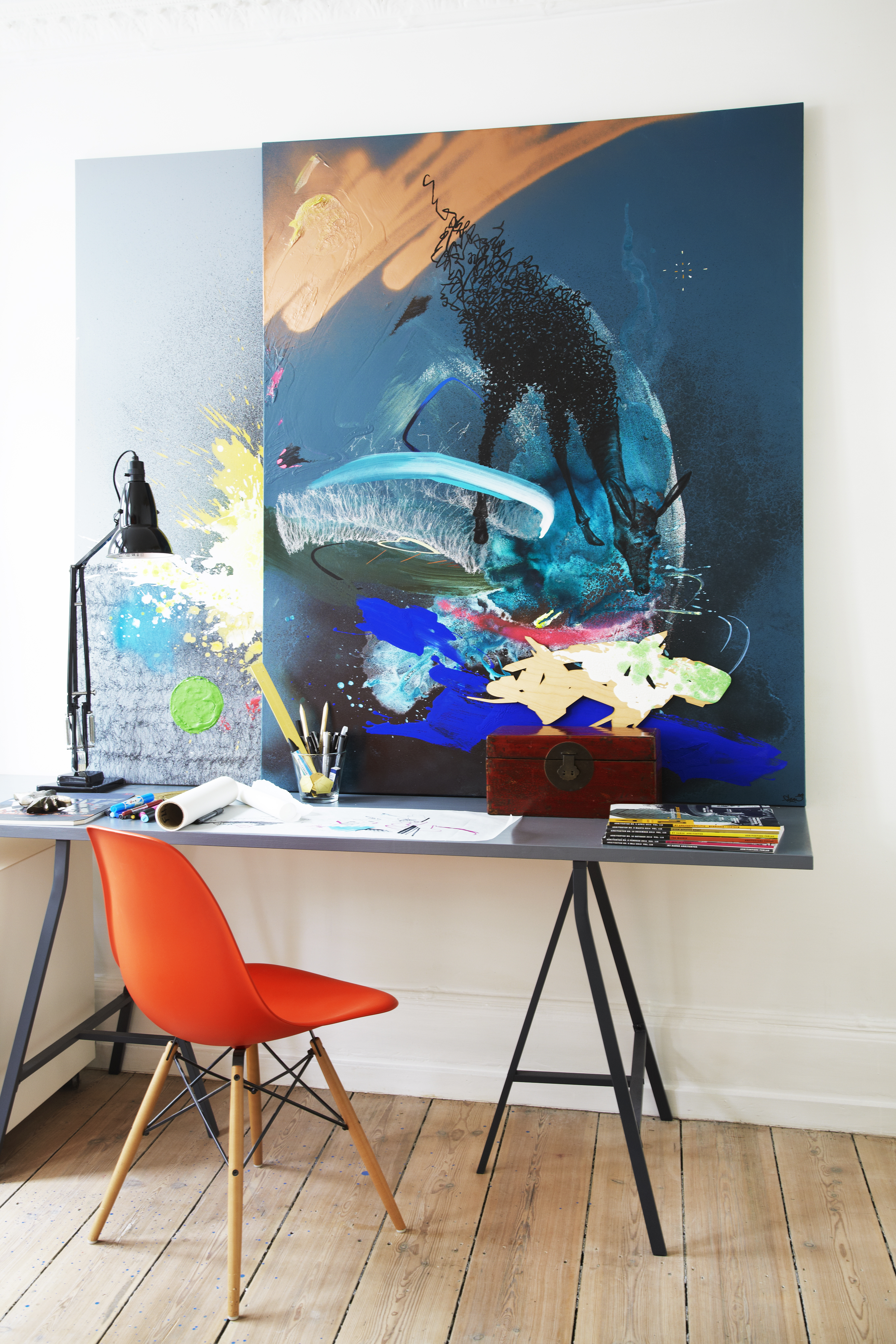 Stort abstrakt maleri i blå nuancer, over skrivebord med rød stol.